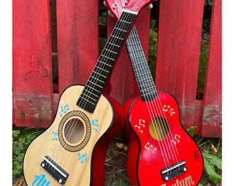 May include: Two wooden ukuleles, one with a natural light wood finish and the other painted bright red. Both feature black fretboards, tuning pegs, and musical note graphics. The natural ukulele has the name Atlas, and the red one has Taytum.