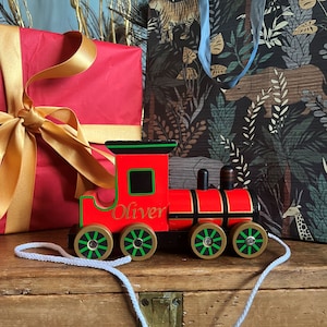 May include: A red wooden toy train with black accents and gold wheels. The train has the name "Oliver" printed on the side. The train is on a wooden surface with a white string attached to it.