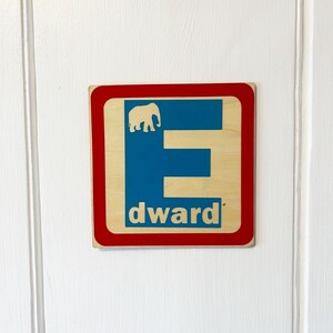 Könnte beinhalten: Ein Holzschild mit rotem Rand und blauem Hintergrund. Der Buchstabe "E" ist in Weiß auf dem blauen Hintergrund gedruckt. Eine Elefanten-Silhouette ist über dem Buchstaben gedruckt. Das Wort "dward" ist unter dem Buchstaben gedruckt.