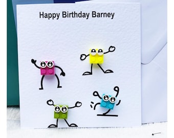 Op de afbeelding: Een vierkante witte verjaardagskaart met de tekst Happy Birthday Barney in het zwart gedrukt. De kaart bevat vier kleurrijke plastic bouwsteentjes die op het papier zijn bevestigd, elk met armen, benen en ogen getekend in zwarte inkt.
