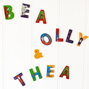 以下が含まれることがあります： 「BEA OLLY & THEA」と綴られたカラフルな木製文字。各文字は異なる色で塗装され、動物のイラストで装飾されています。文字は白い背景に配置されています。