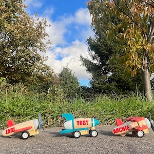 Puede incluir: Tres aviones de juguete de madera con pintura roja, azul y verde. Los aviones tienen nombres: "Carl", "Toby" y "Archie".