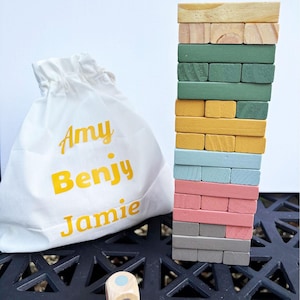 Könnte beinhalten: Ein Holzblock-Stapelspiel mit einem Turm aus farbigen Blöcken in Grün-, Gelb-, Blau-, Rosa- und Grautönen. Neben dem Turm befindet sich ein weißer Beutel mit den Namen "Amy Benjy Jamie" in gelben Buchstaben.
