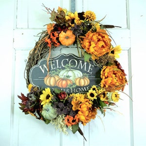 May include: A fall-themed wreath with a welcome sign that says "Welcome to our home". The wreath is made of grapevine and decorated with pumpkins, sunflowers, and other fall foliage.