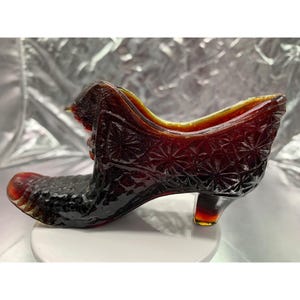 May include: A deep amber-colored glass shoe figurine featuring a textured starburst pattern across the side and a pebbled design on the toe area. The decorative object has a curved silhouette with a small heel and an open top opening.