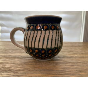 May include: A ceramic mug with a rounded body and a handle. The mug features a dark blue rim and a pattern of vertical white stripes with teal and orange floral accents. The mug sits on a wooden surface.