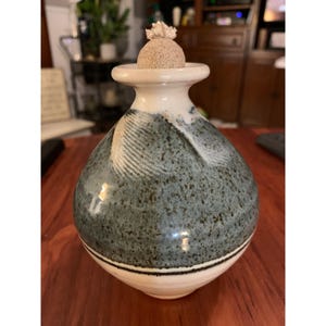 May include: A ceramic vase with a speckled blue and white glaze. The vase has a round body and a narrow neck with a white rim. The top of the vase has a small, round, beige lid.