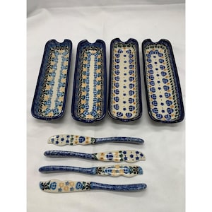 May include: Four rectangular ceramic serving dishes and four matching spreaders, each decorated with a blue, yellow, and green floral pattern on a cream background. The dishes feature scalloped edges and deep blue rims, while the spreaders have patterned handles and blades.