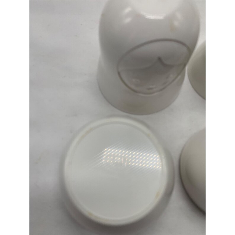 Puede incluir: Un conjunto de mu&ntilde;ecas rusas de cer&aacute;mica blanca con superficies lisas y redondeadas. Una mu&ntilde;eca se muestra desde arriba, revelando una base circular con un logotipo tenue en relieve. Las otras mu&ntilde;ecas son parcialmente visibles con contornos faciales.