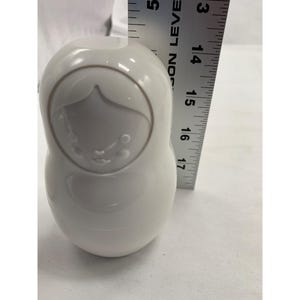 Puede incluir: Una figura de mu&ntilde;eca rusa de cer&aacute;mica blanca lisa y brillante con un dise&ntilde;o de cara en relieve minimalista. Una regla de metal est&aacute; colocada verticalmente junto al objeto, indicando una altura de unos 11,5 cent&iacute;metros para el cuerpo.