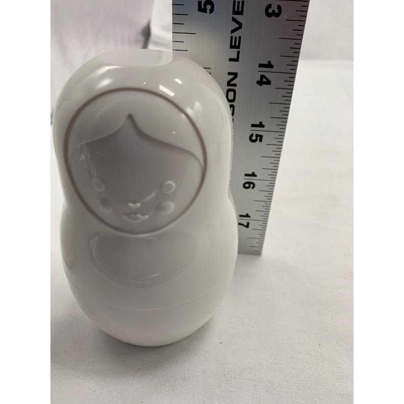 Puede incluir: Una figura de mu&ntilde;eca rusa de cer&aacute;mica blanca lisa y brillante con un dise&ntilde;o de cara en relieve minimalista. Una regla de metal est&aacute; colocada verticalmente junto al objeto, indicando una altura de unos 11,5 cent&iacute;metros para el cuerpo.