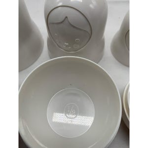 Puede incluir: Un juego de tazas medidoras encajables de color blanco roto con forma de mu&ntilde;ecas. La taza frontal est&aacute; abierta, revelando un interior circular con la fracci&oacute;n 1/2 en relieve en el centro. Las otras tazas est&aacute;n cerradas y muestran un rostro.