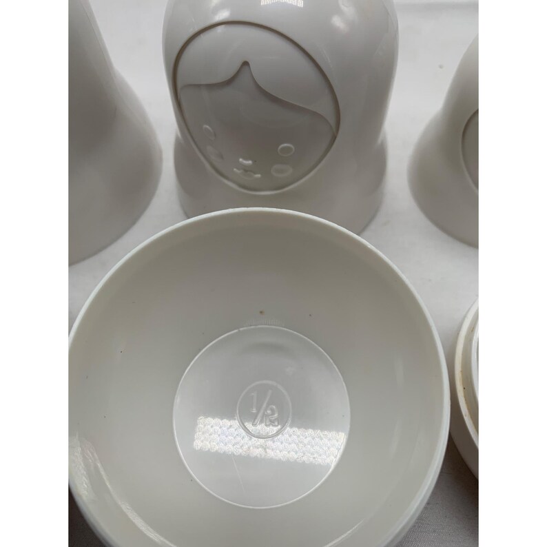 Puede incluir: Un juego de tazas medidoras encajables de color blanco roto con forma de mu&ntilde;ecas. La taza frontal est&aacute; abierta, revelando un interior circular con la fracci&oacute;n 1/2 en relieve en el centro. Las otras tazas est&aacute;n cerradas y muestran un rostro.