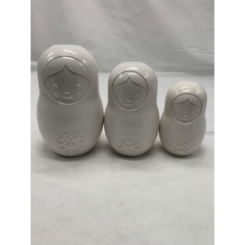 Puede incluir: Tres mu&ntilde;ecas rusas blancas s&oacute;lidas dispuestas en fila de mayor a menor. Cada mu&ntilde;eca presenta un acabado liso y brillante con un rostro moldeado y un dise&ntilde;o floral en relieve &uacute;nico en la parte inferior del cuerpo de cada figura.