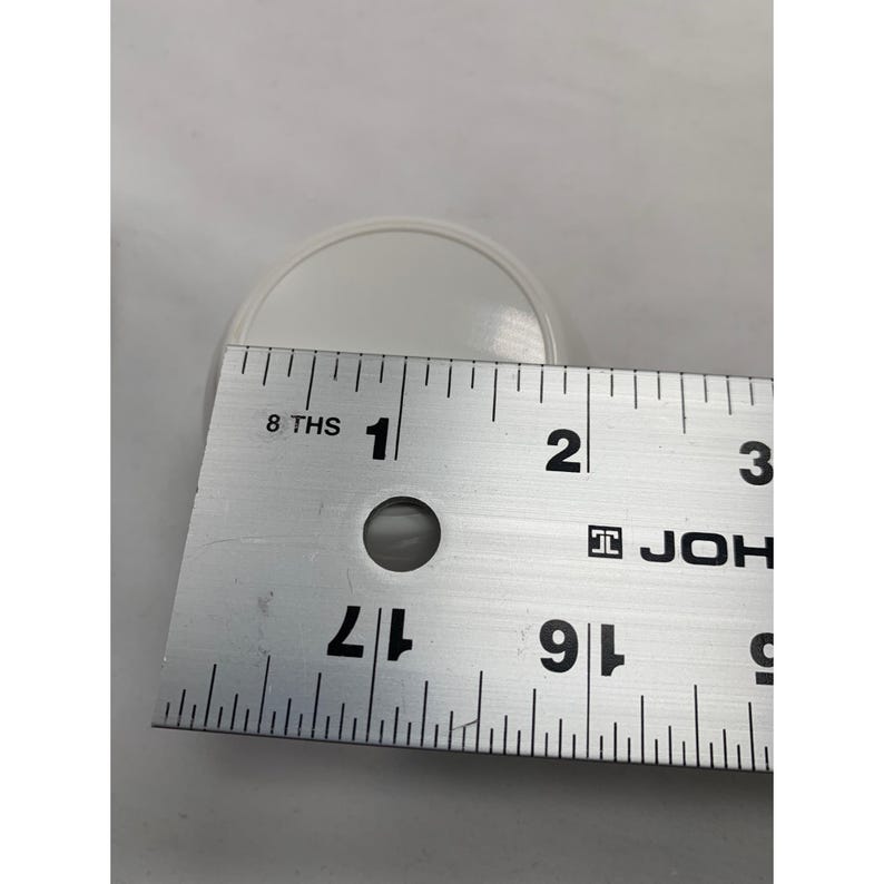 Puede incluir: Un objeto circular blanco colocado detr&aacute;s de una regla de metal. La regla muestra que el objeto tiene un di&aacute;metro de aproximadamente 5,7 cent&iacute;metros. La regla presenta marcas negras, el texto 8 THS y la marca JOHN en su superficie.