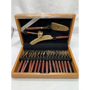 May include: A wooden storage box with a black interior containing a set of brass flatware with dark wood handles. The set includes a cake server, a small ladle, a curved knife, and twelve forks, all arranged in custom slots within the box.