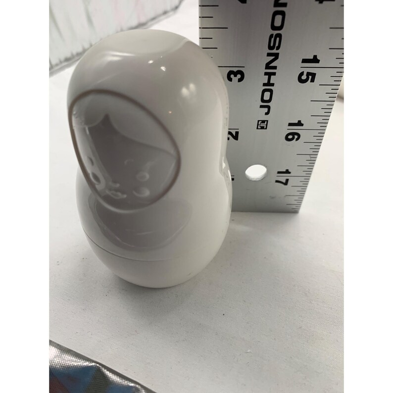 Puede incluir: Una figura de mu&ntilde;eca rusa blanca, lisa y brillante, con un contorno facial moldeado. Una regla met&aacute;lica est&aacute; colocada verticalmente junto al objeto, indicando una altura de aproximadamente 8,9 cm desde la base hasta la parte superior.