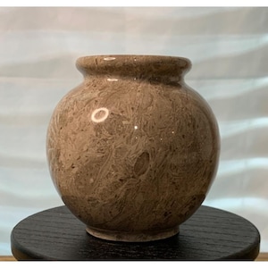 May include: A brown, spherical vase with a textured surface. The vase has a rounded body and a small, cylindrical neck. The color is a mix of light and dark brown, with a marbled appearance. The vase is sitting on a black, round stand.