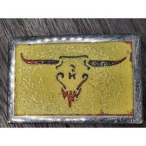 May include: Rectangular silver-framed belt buckle with a yellow background. The buckle features a stylized bull skull design with red and black horns and a red lower jaw. The design is centered on the buckle.