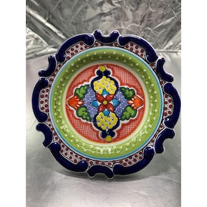 May include: A decorative ceramic plate with a vibrant, hand-painted design. The plate features a central floral motif in red, yellow, blue, and green, surrounded by a green border with white dots. The rim is dark blue with a scalloped edge.