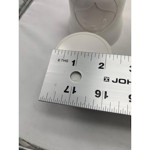 Puede incluir: Un recipiente de cer&aacute;mica blanca con un dise&ntilde;o de rostro moldeado en la parte superior. Una regla de metal est&aacute; colocada sobre la parte superior, mostrando un di&aacute;metro de aproximadamente 6,35 cm. El objeto tiene un acabado liso y mate.