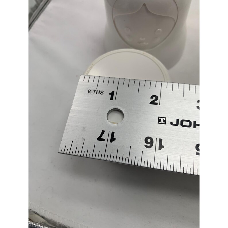 Puede incluir: Un recipiente de cer&aacute;mica blanca con un dise&ntilde;o de rostro moldeado en la parte superior. Una regla de metal est&aacute; colocada sobre la parte superior, mostrando un di&aacute;metro de aproximadamente 6,35 cm. El objeto tiene un acabado liso y mate.