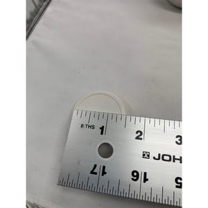 Puede incluir: Una peque&ntilde;a tapa o tap&oacute;n redondo de pl&aacute;stico color blanco hueso colocado detr&aacute;s de una regla met&aacute;lica. La regla muestra que el objeto tiene un di&aacute;metro de aproximadamente 3,2 cent&iacute;metros. El objeto presenta una superficie superior circular lisa con un borde ligeramente elevado.