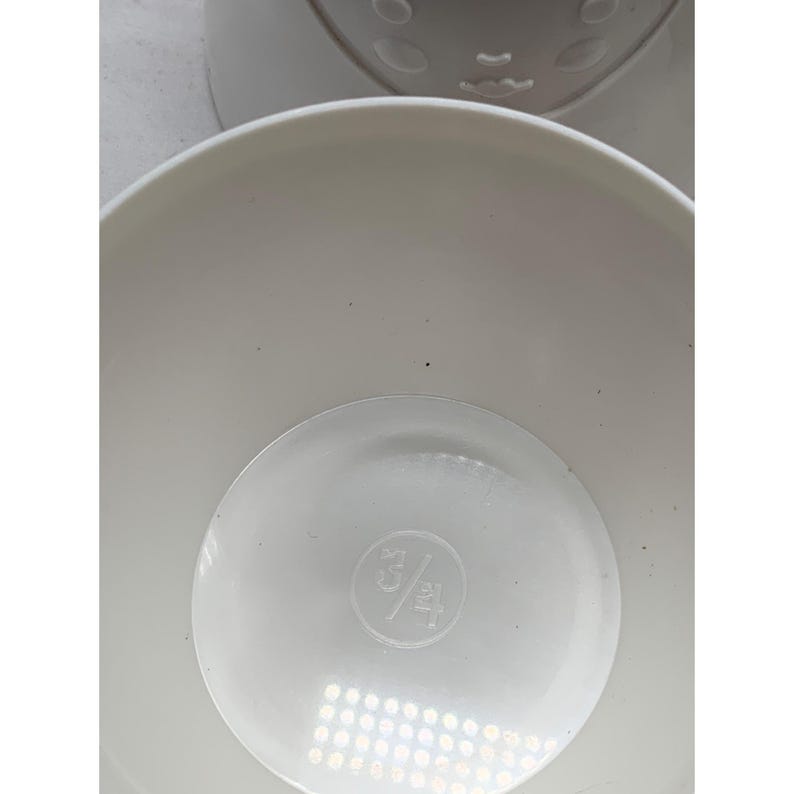Puede incluir: Una taza medidora de pl&aacute;stico blanco con un interior liso y redondeado. El fondo de la taza presenta una marca de medici&oacute;n en relieve de 3/4 cup, centrada dentro de una hendidura circular en la base del recipiente.