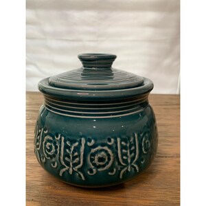 May include: Teal ceramic jar with a lid. The jar has a glossy finish and is decorated with embossed floral and circular patterns. The lid has a small knob and concentric rings. The jar sits on a wooden surface.