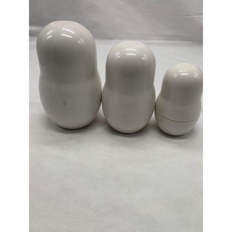 Puede incluir: Tres mu&ntilde;ecas rusas blancas, lisas y brillantes, de diferentes tama&ntilde;os, dispuestas en fila. Cada mu&ntilde;eca presenta una forma redondeada y bulbosa con una costura horizontal que separa las mitades superior e inferior, de aspecto uniforme.
