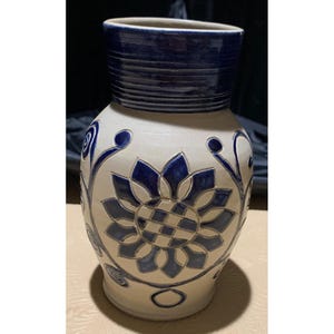May include: A ceramic vase with a cream-colored body and a dark blue rim. The vase features a large blue floral design on the front and smaller blue floral accents. The vase is approximately 10 inches tall.