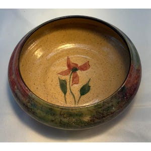 May include: A round ceramic bowl with a speckled tan interior and a painted red flower with green leaves. The rim features bands of red, green, and brown. This decorative bowl is suitable for serving or display.