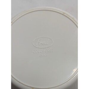 Puede incluir: Un objeto circular de pl&aacute;stico blanco con un borde elevado. El centro presenta el texto en relieve "Fred" dentro de un &oacute;valo, seguido del texto "&copy;2009 FRED" y "CHINA" impreso en una fuente clara y legible sobre la superficie blanca lisa.