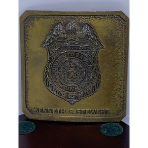 May include: A rectangular brass plaque with a detailed emblem featuring an eagle and the text "Alcohol, Tobacco, Firearms, and Explosives." The plaque also includes the name "Kenneth F. Stewart."