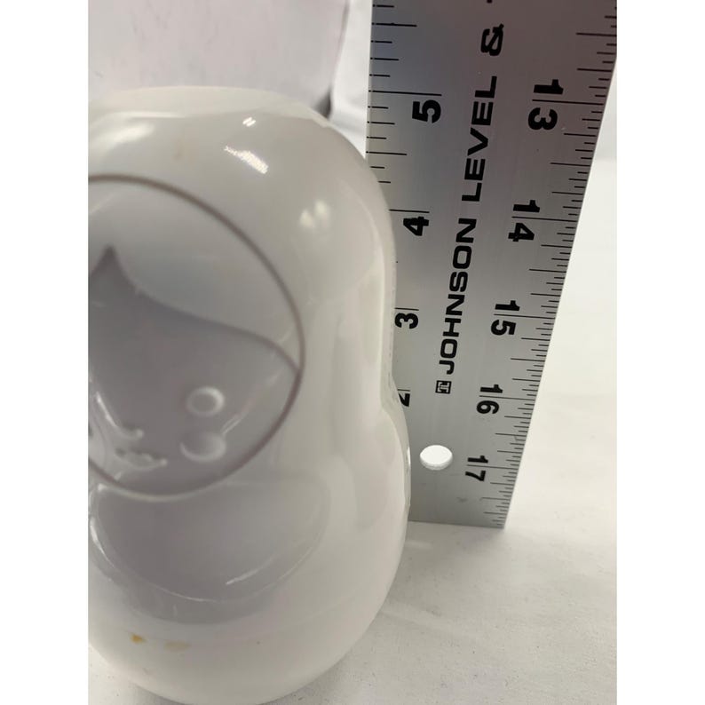 Puede incluir: Una figura blanca, lisa y redondeada con forma de mu&ntilde;eca rusa, que presenta rasgos faciales en relieve y un contorno de cabello. Una regla de metal est&aacute; colocada verticalmente junto al objeto, mostrando una altura de unos 14 cm.