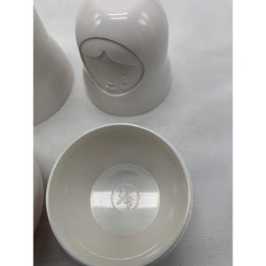 Puede incluir: Un conjunto de mu&ntilde;ecas rusas de pl&aacute;stico blanco con un acabado liso. Una mu&ntilde;eca est&aacute; cerrada, mostrando un rostro moldeado, mientras que otra est&aacute; abierta, revelando la base interior con el texto 1/4 cup grabado en el fondo del recipiente.