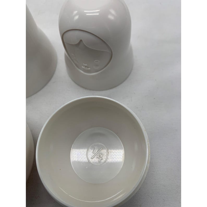 Puede incluir: Un conjunto de mu&ntilde;ecas rusas de pl&aacute;stico blanco con un acabado liso. Una mu&ntilde;eca est&aacute; cerrada, mostrando un rostro moldeado, mientras que otra est&aacute; abierta, revelando la base interior con el texto 1/4 cup grabado en el fondo del recipiente.
