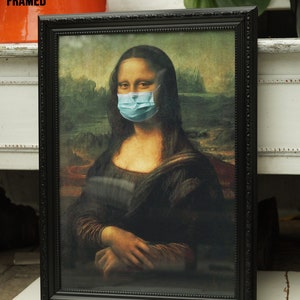 Puede incluir: Una impresi&oacute;n enmarcada en negro de la Mona Lisa con una m&aacute;scara quir&uacute;rgica azul. La impresi&oacute;n est&aacute; en un marco negro adornado.