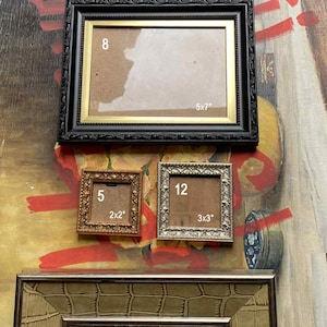 May include: A selection of decorative picture frames in different sizes and designs. The frames include a black frame with gold trim (12.7x17.8 cm), a small gold frame (5.1x5.1 cm), a silver frame (7.6x7.6 cm), and a brown frame with a reptile skin pattern.