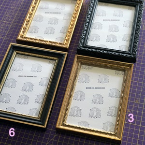 Op de afbeelding: Vier fotolijsten in verschillende stijlen en kleuren, waaronder goud en zwart. De lijsten zijn in een rij op een paarse rooster-oppervlak gerangschikt. De lijsten zijn allemaal 4x6 inch en bevatten een afdruk van een regenboog met sterren.
