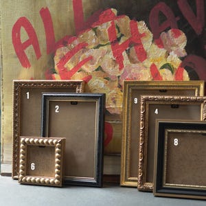 May include: A selection of rectangular picture frames in gold, black, and brown. The frames vary in size and design, with both ornate and simple styles. The background features a painting with red text and floral imagery.