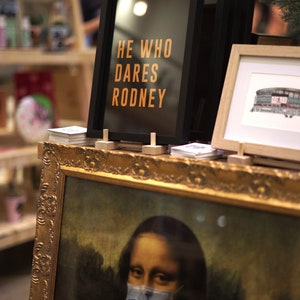 Puede incluir: Una impresi&oacute;n enmarcada con el texto "He who dares Rodney" en letras naranjas sobre fondo negro. La impresi&oacute;n est&aacute; sentada en un estante con otras impresiones enmarcadas y una pintura enmarcada en oro de la Mona Lisa con una m&aacute;scara facial azul.