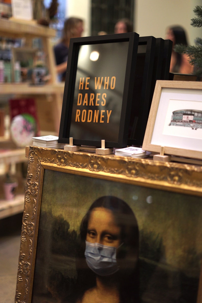 Puede incluir: Una impresi&oacute;n enmarcada con el texto "He who dares Rodney" en letras naranjas sobre fondo negro. La impresi&oacute;n est&aacute; sentada en un estante con otras impresiones enmarcadas y una pintura enmarcada en oro de la Mona Lisa con una m&aacute;scara facial azul.