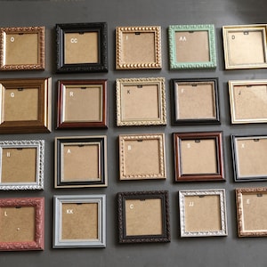 May include: Twenty square picture frames in a variety of colours and designs. Frames include gold, black, brown, silver, and pink, with ornate and simple styles. Each frame has a neutral backing and a letter label.