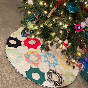 May include: A colorful, quilted Christmas tree skirt with a patchwork design. The skirt features various patterned circles in red, green, blue, yellow, and black and white, with a black and white rope trim. The skirt is placed under a decorated Christmas tree.