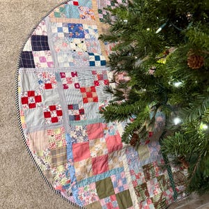 May include: A circular patchwork tree skirt featuring a variety of multi-colored fabric squares in shades of red, blue, pink, green, and tan. The quilt-style design is finished with a black and white striped fabric border along the outer edge.