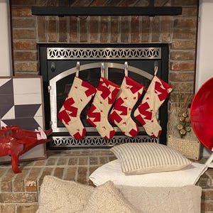 May include: Four cream-colored Christmas stockings with a repeating red geometric pattern hanging from a fireplace mantel. Each stocking features a red cuff and toe section, suspended by a loop from a silver metal hook on the fireplace screen.