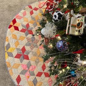 Könnte beinhalten: Ein handgefertigter Weihnachtsbaumrock mit Patchwork-Design. Der Rock zeigt verschiedene Stoffe in Rot, Rosa, Gelb und Creme, angeordnet in einem Sternmuster. Der Rand ist schwarz-weiß gestreift. Der Rock liegt um einen geschmückten Weihnachtsbaum.