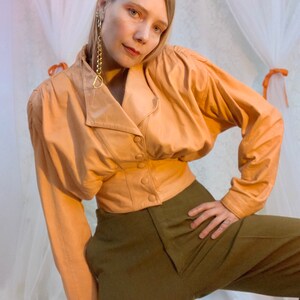 May include: A peach-colored leather jacket with a cinched waist and long sleeves. The jacket has a button-up front and a collar. The model is wearing the jacket with olive green pants.