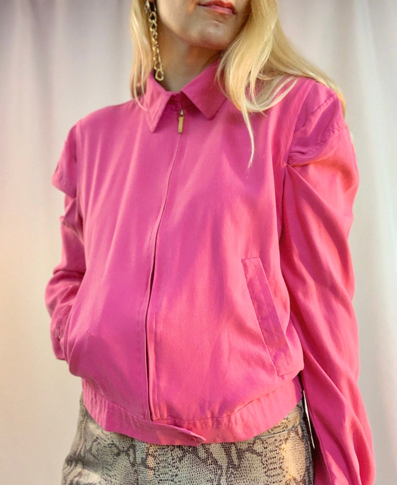May include: A pink cropped jacket with a zipper closure and a collared neckline. The jacket has puffed sleeves and a single pocket on the left side.